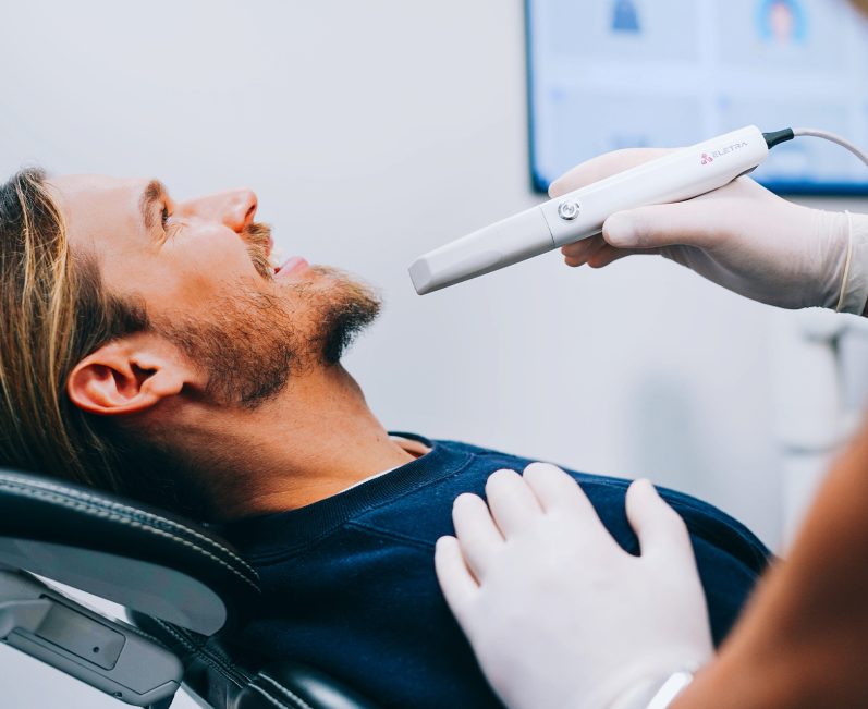 Mann sitzt im Zahnarztstuhl, während ein Zahnarzt mit einem Intraoralscanner arbeitet.